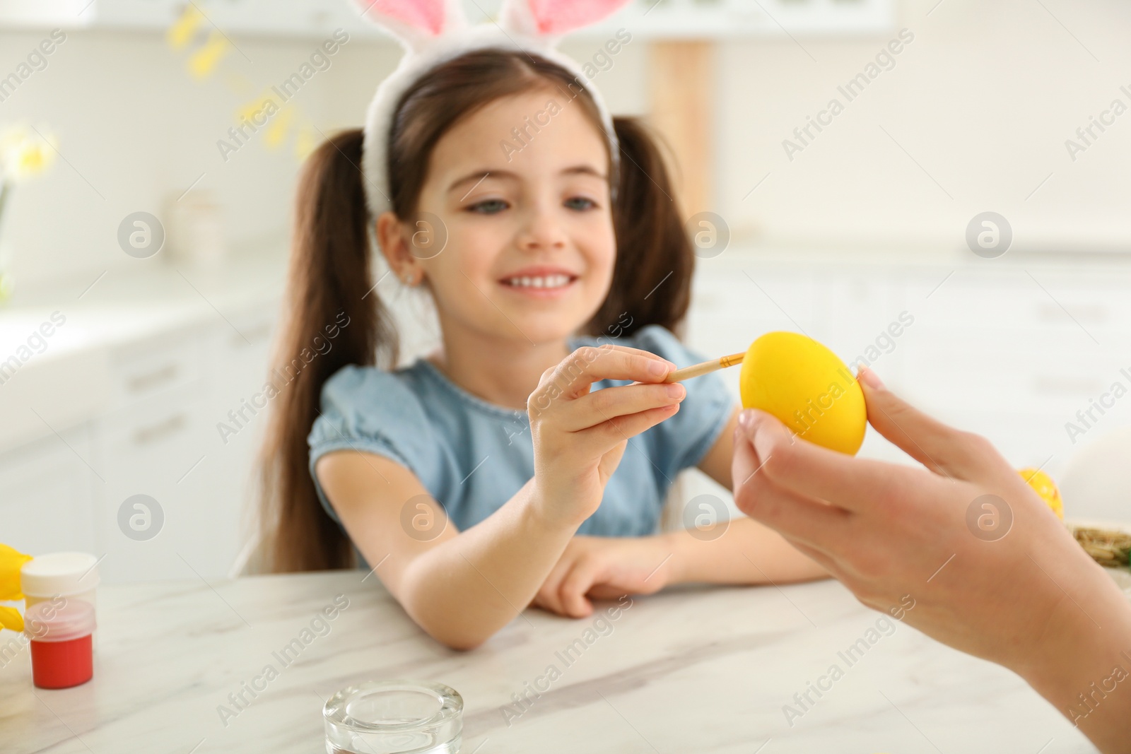 Photo of Happy little girl with bunny ears headband and her mother painting Easter egg in kitchen