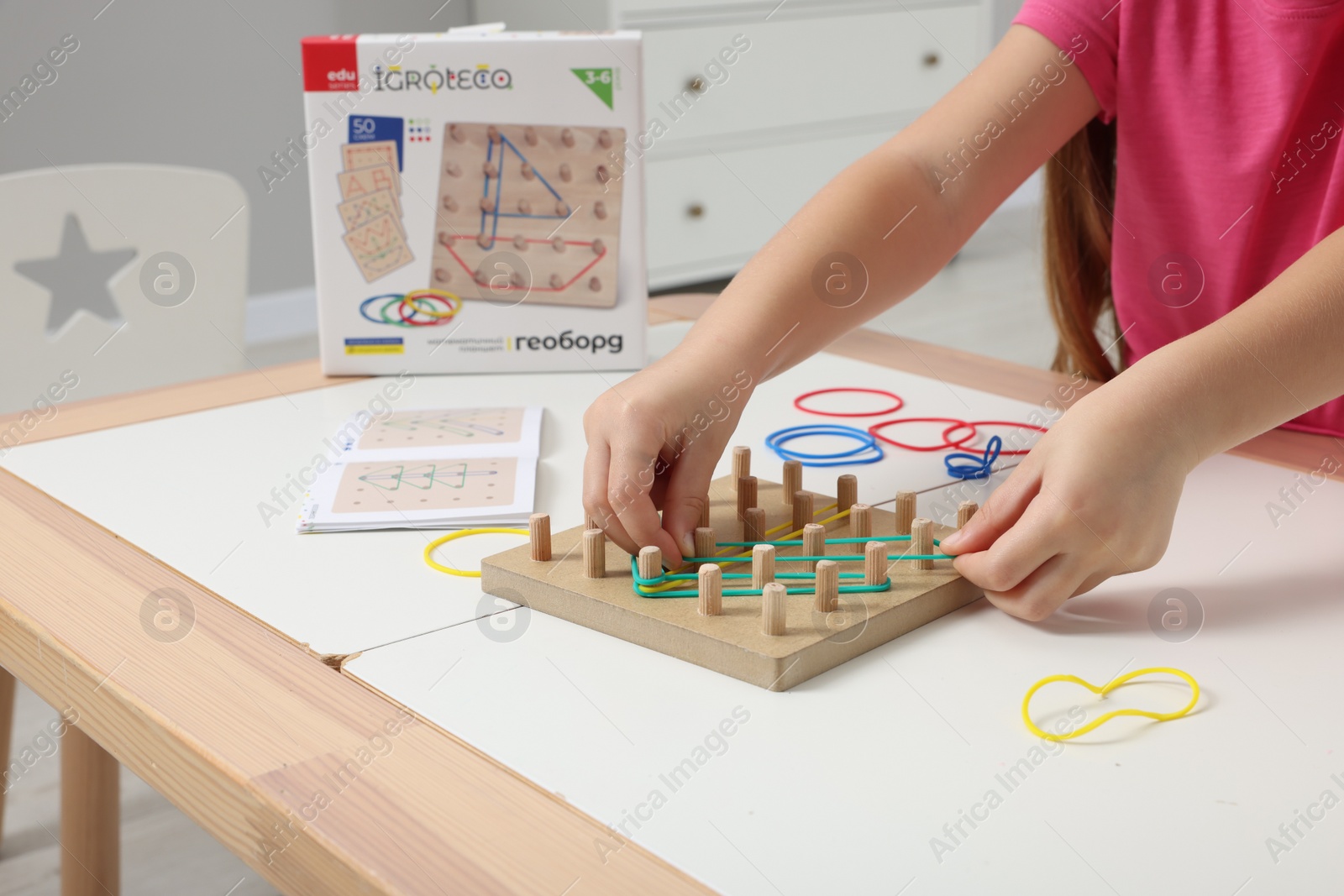 Photo of Motor skills development. Girl playing with geoboard and rubber bands at white table indoors, closeup