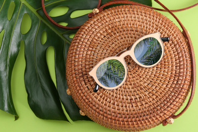 Bamboo bag and sunglasses with reflection of palm trees on green background, flat lay 