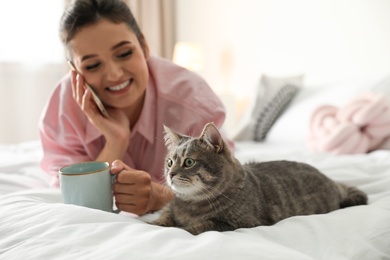 Photo of Young woman with cup of coffee talking on phone while lying near cute cat in bedroom. Pet and owner