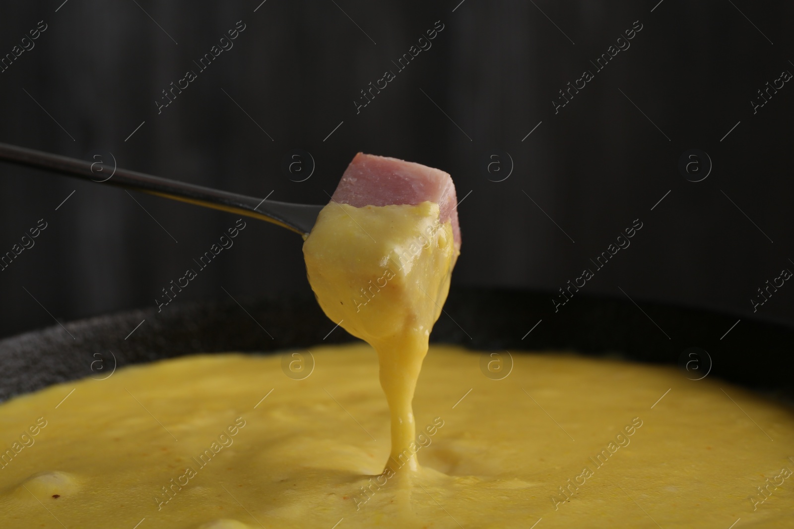 Photo of Dipping piece of ham into fondue pot with tasty melted cheese against dark gray background, closeup