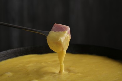 Photo of Dipping piece of ham into fondue pot with tasty melted cheese against dark gray background, closeup