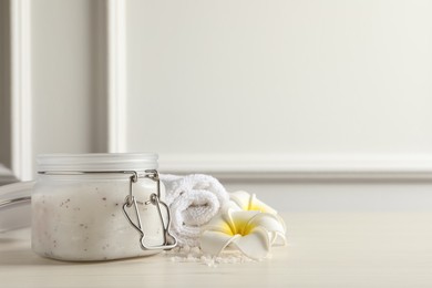 Photo of Body scrub in glass jar, towel and plumeria flowers on white wooden table, space for text