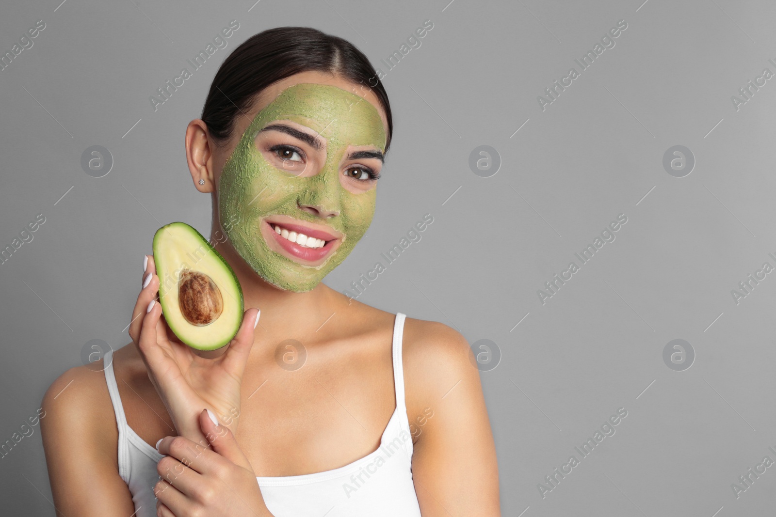 Photo of Young woman with clay mask on her face holding avocado against grey background, space for text. Skin care