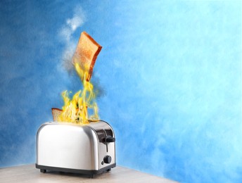 Toaster flaming up while cooking slices of bread on white wooden table against blue background. Unsafe appliance 