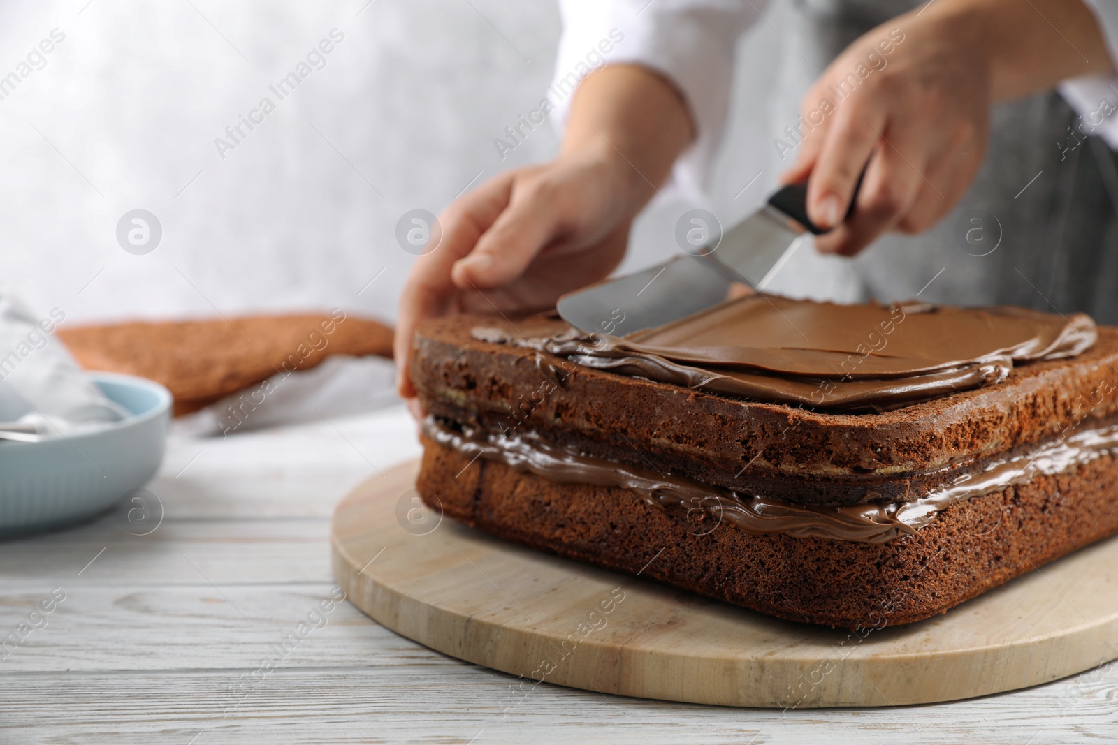 Photo of Woman smearing sponge cake with chocolate cream at white wooden table, closeup