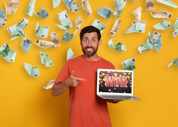 Betting. Happy man pointing at laptop with word Win! under money shower on yellow background
