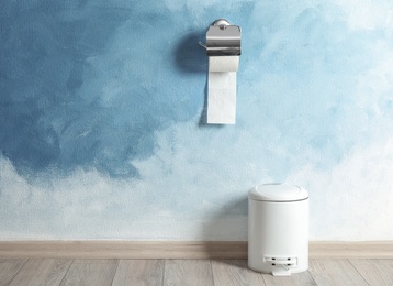 Photo of Toilet paper holder with roll and trash bin near color wall. Space for text