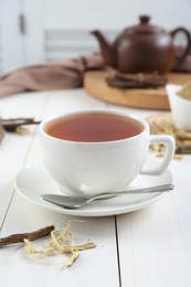 Photo of Aromatic licorice tea in cup and dried sticks of licorice root on white wooden table