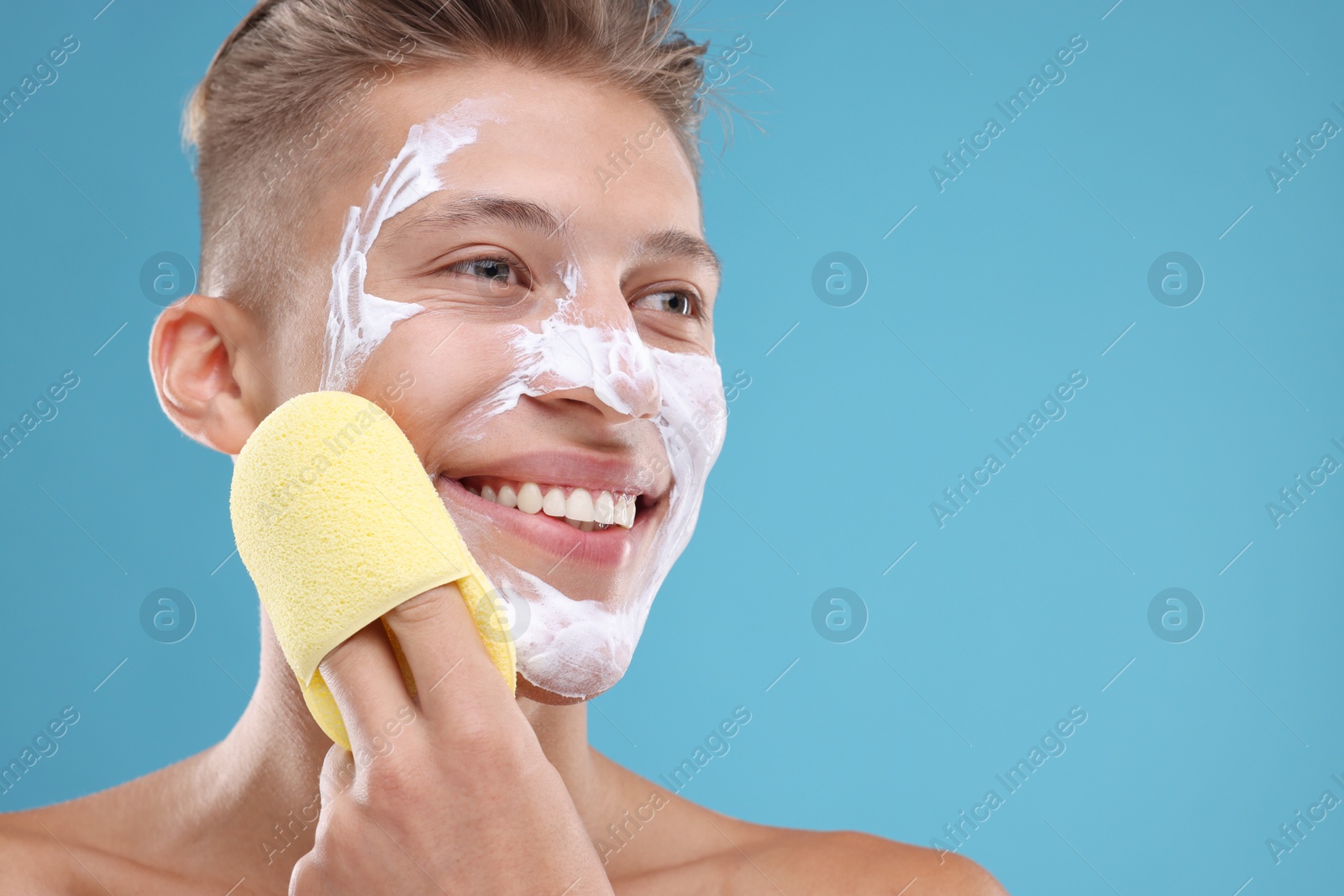 Photo of Happy young man washing off face mask with sponge on light blue background. Space for text
