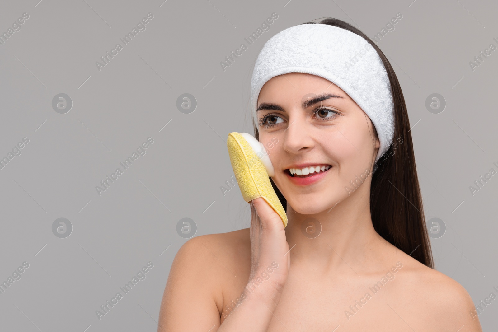 Photo of Young woman with headband washing her face using sponge on light grey background, space for text