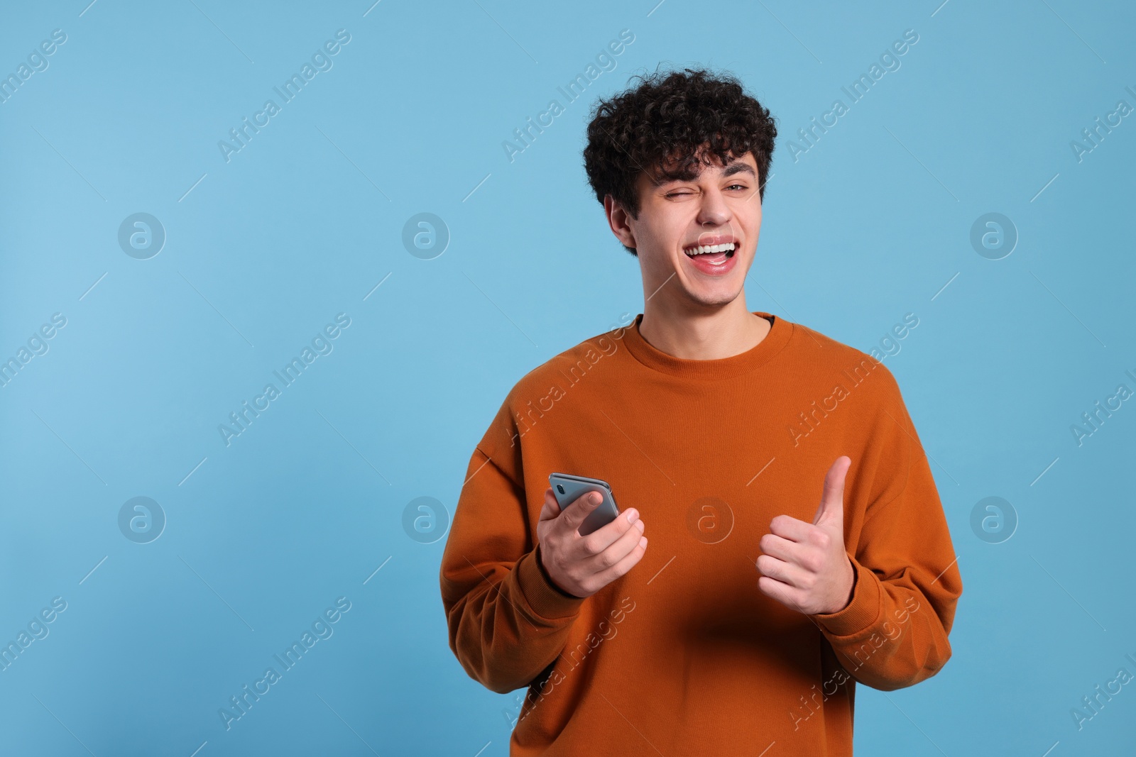 Photo of Handsome young man with smartphone showing thumb up on light blue background. Space for text