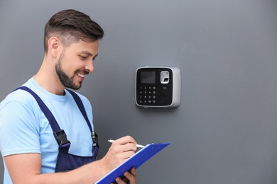 Photo of Male technician with clipboard near installed alarm system indoors