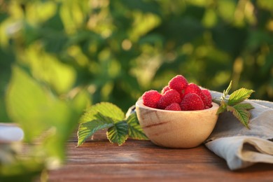 Photo of Tasty ripe raspberries in bowl and green leaves on wooden table outdoors, space for text