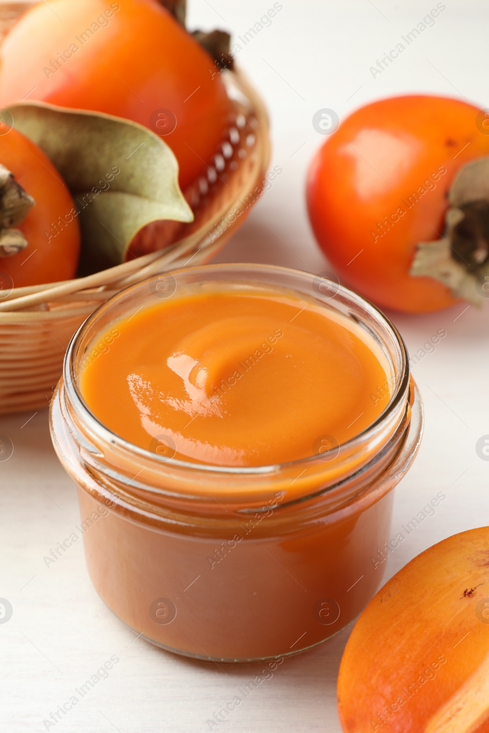 Photo of Delicious persimmon jam in glass jar and fresh fruits on white wooden table, closeup