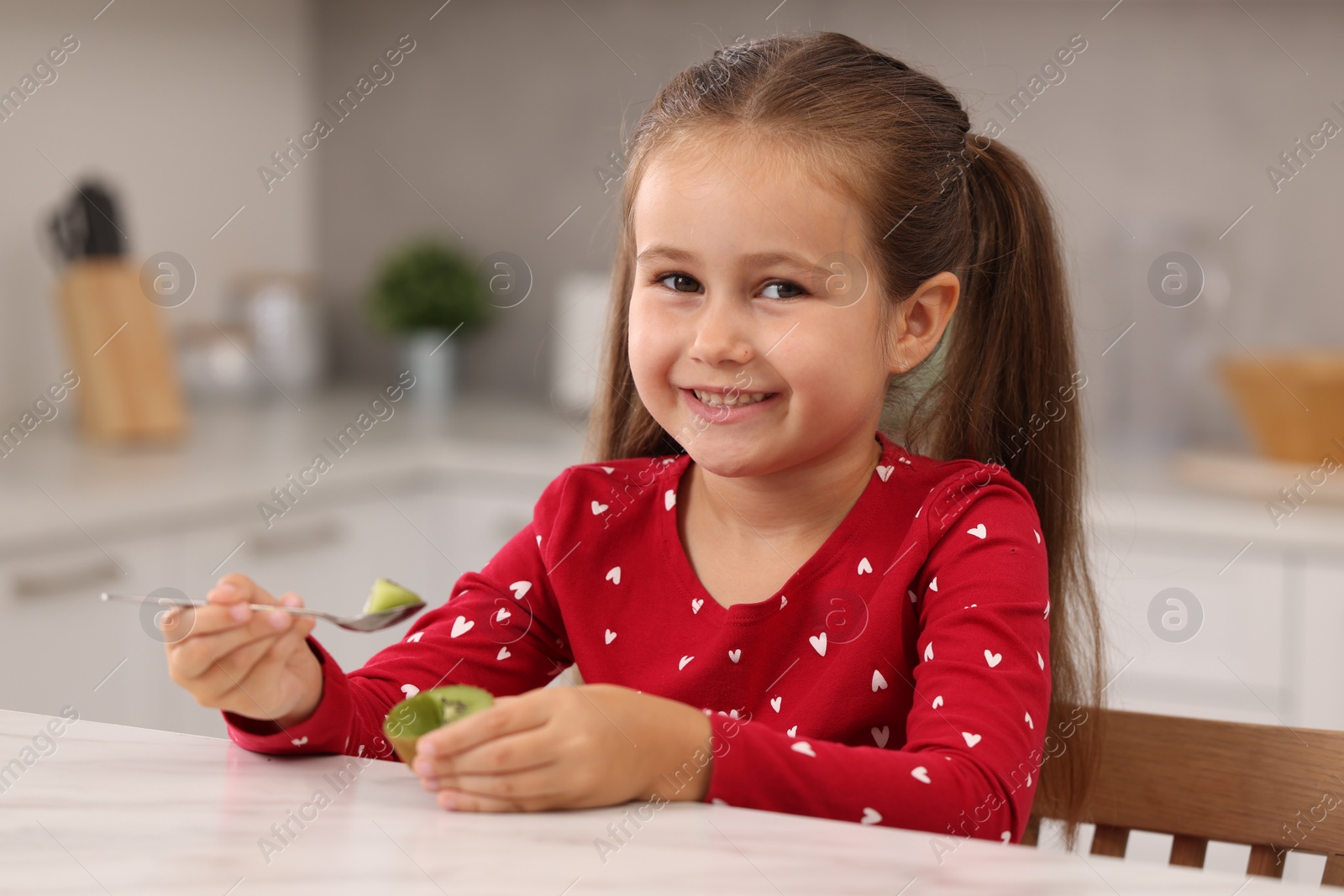 Photo of Happy girl eating kiwi at white table in kitchen