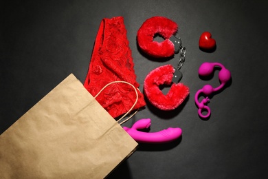 Photo of Paper shopping bag with different sex toys and underwear on black background, flat lay