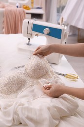 Dressmaker creating beautiful wedding dress with embroidered bustline and bow at table in workshop, closeup