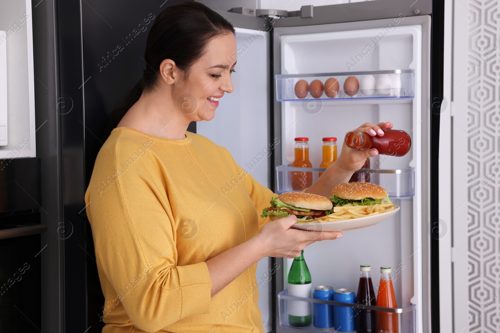 Photo of Happy overweight woman with ketchup and burgers near fridge in kitchen