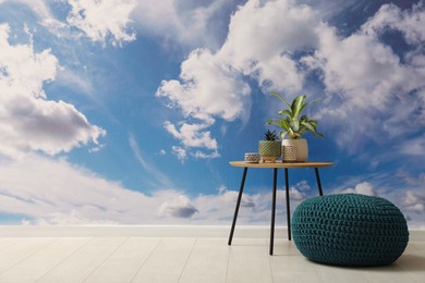 Blue sky with clouds as wallpaper pattern in room. Side table with houseplants and knitted pouf near wall