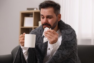 Sick man wrapped in blanket with tissue and cup of drink on sofa at home. Cold symptoms