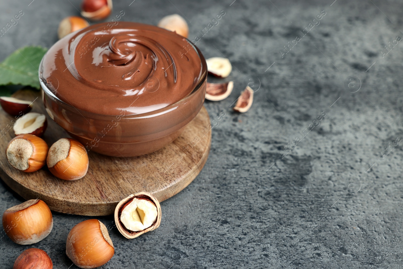 Photo of Glass bowl with tasty chocolate hazelnut spread and nuts on grey table. Space for text