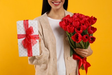 Photo of Happy woman with red tulip bouquet and gift box on yellow background, closeup. 8th of March celebration