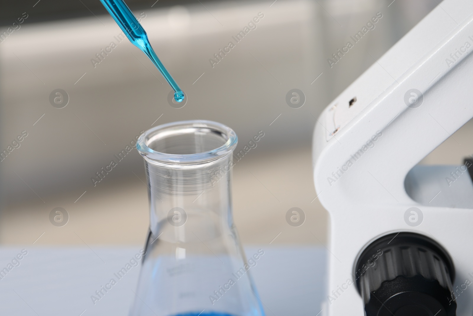 Photo of Laboratory analysis. Dripping blue liquid into flask near microscope on table, closeup