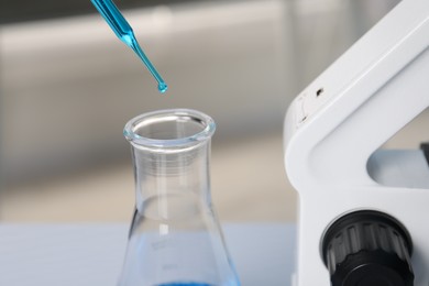 Laboratory analysis. Dripping blue liquid into flask near microscope on table, closeup