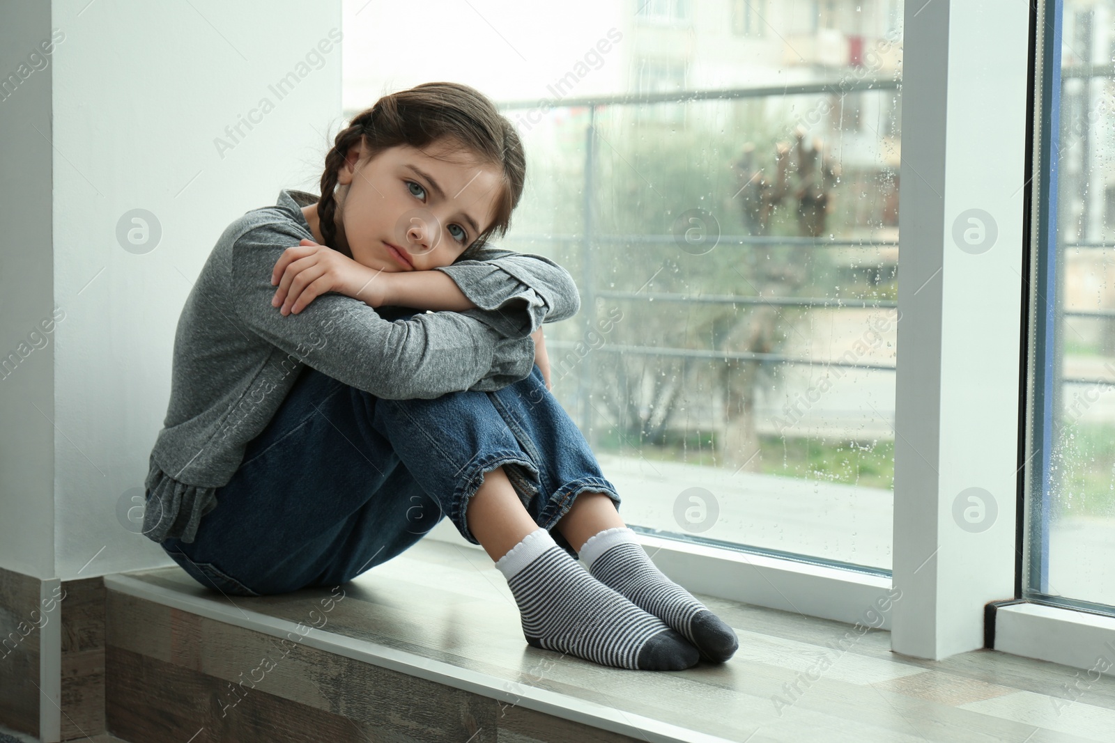 Photo of Sad little girl sitting on window sill indoors