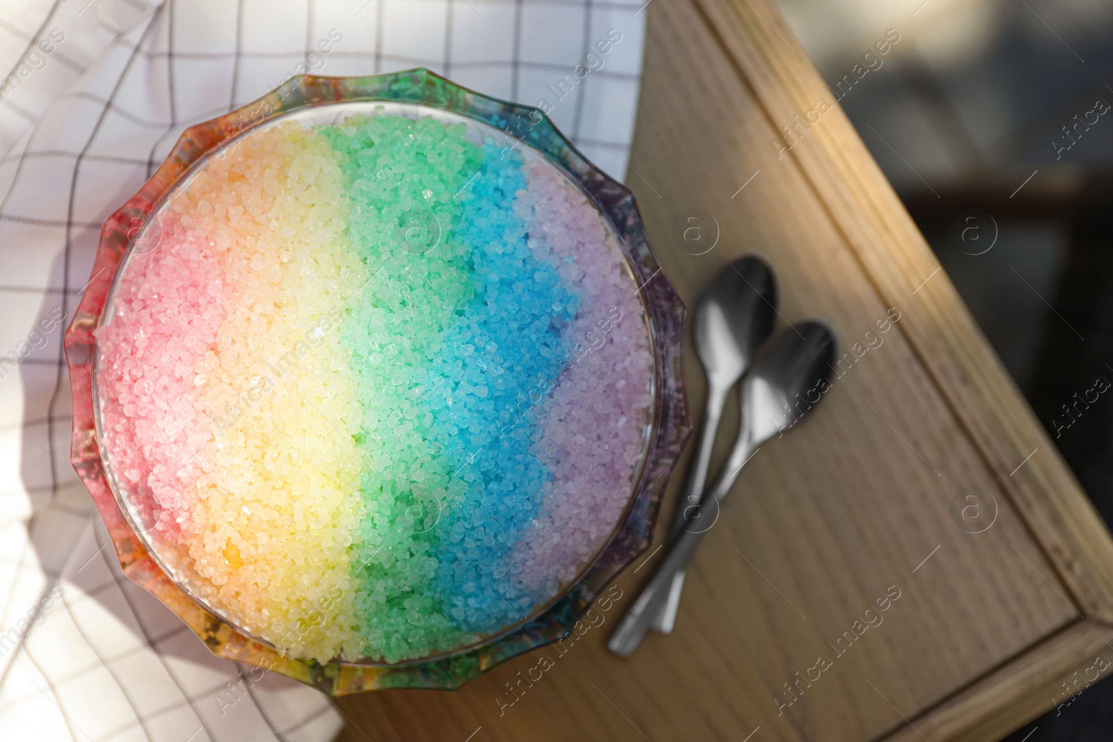 Photo of Rainbow shaving ice in glass dessert bowl and spoons on wooden table, flat lay