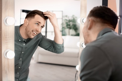 Photo of Young man with trendy hairstyle looking in mirror indoors