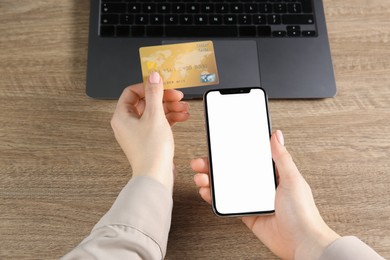 Online payment. Woman using credit card and smartphone with blank screen near laptop at wooden table, closeup