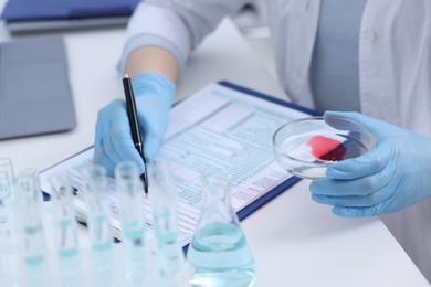 Laboratory worker holding petri dish with blood sample while working at white table, closeup