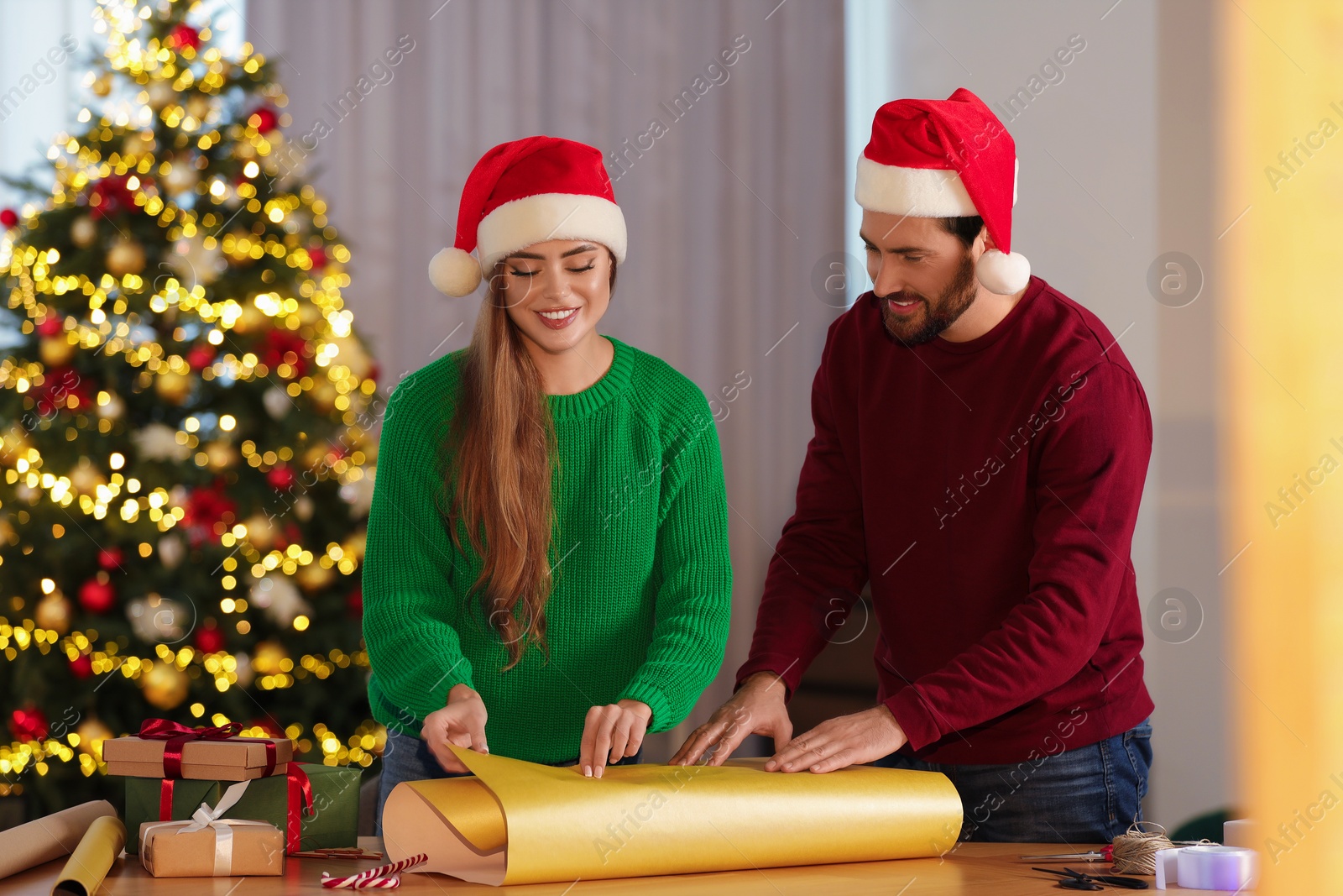 Photo of Happy couple in Santa hats decorating Christmas gift with wrapping paper at table in room