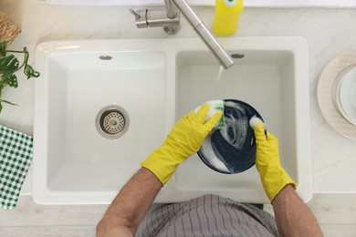 Man in protective gloves washing plate above sink indoors, top view