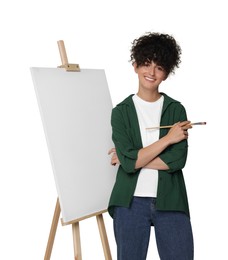 Young woman holding brush near easel with canvas against white background