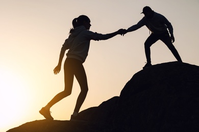 Photo of Silhouettes of man and woman helping each other to climb on hill against sunset