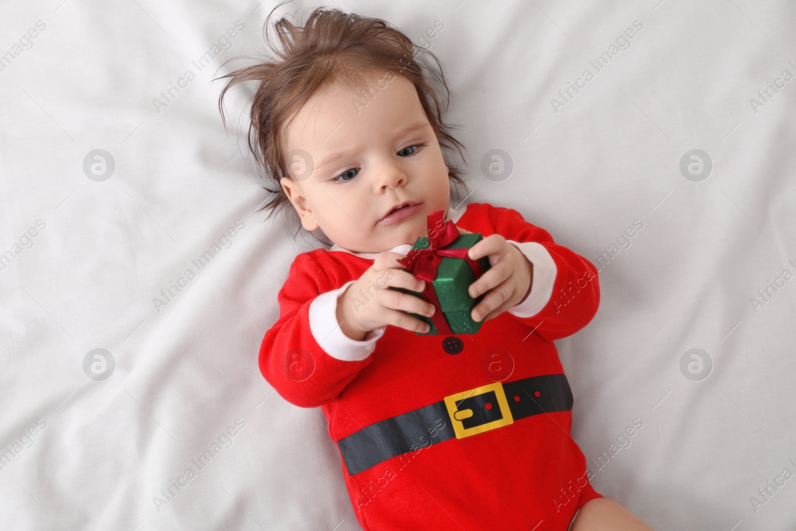 Photo of Cute baby wearing festive Christmas costume with gift box on white bedsheet, top view