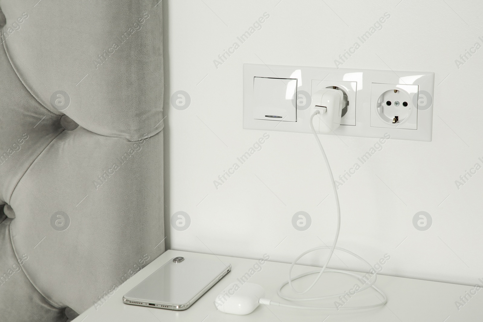 Photo of Charging case of modern wireless earphones plugged into power socket and smartphone on white table indoors