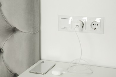 Photo of Charging case of modern wireless earphones plugged into power socket and smartphone on white table indoors