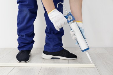 Photo of Man using caulking gun while installing plinth on laminated floor in room, closeup