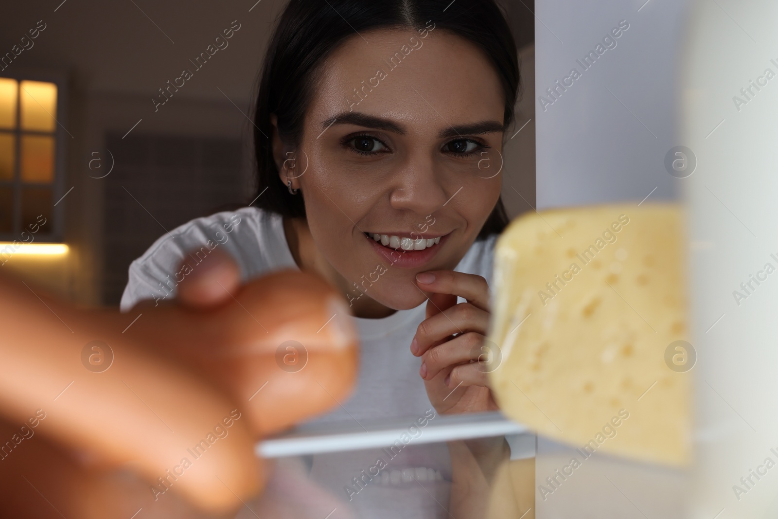 Photo of Young woman taking sausages out of refrigerator in kitchen at night, view from inside