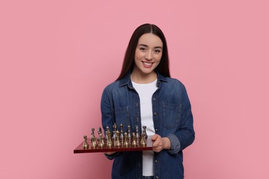 Photo of Happy woman holding chessboard with game pieces on pink background