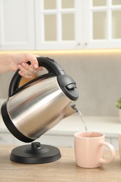 Photo of Woman pouring hot water from electric kettle into cup in kitchen, closeup