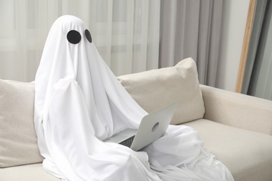 Photo of Creepy ghost. Person covered with white sheet using laptop on sofa at home