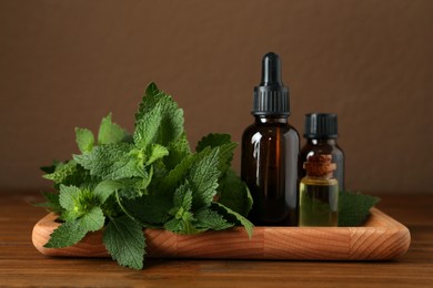 Photo of Tray with bottles of nettle oil and fresh leaves on wooden table against brown background