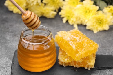 Photo of Pouring sweet golden honey from dipper into jar and pieces of honeycomb on grey table