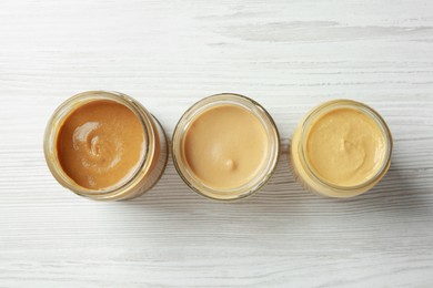 Different types of spicy mustard in glass jars on white wooden table, flat lay