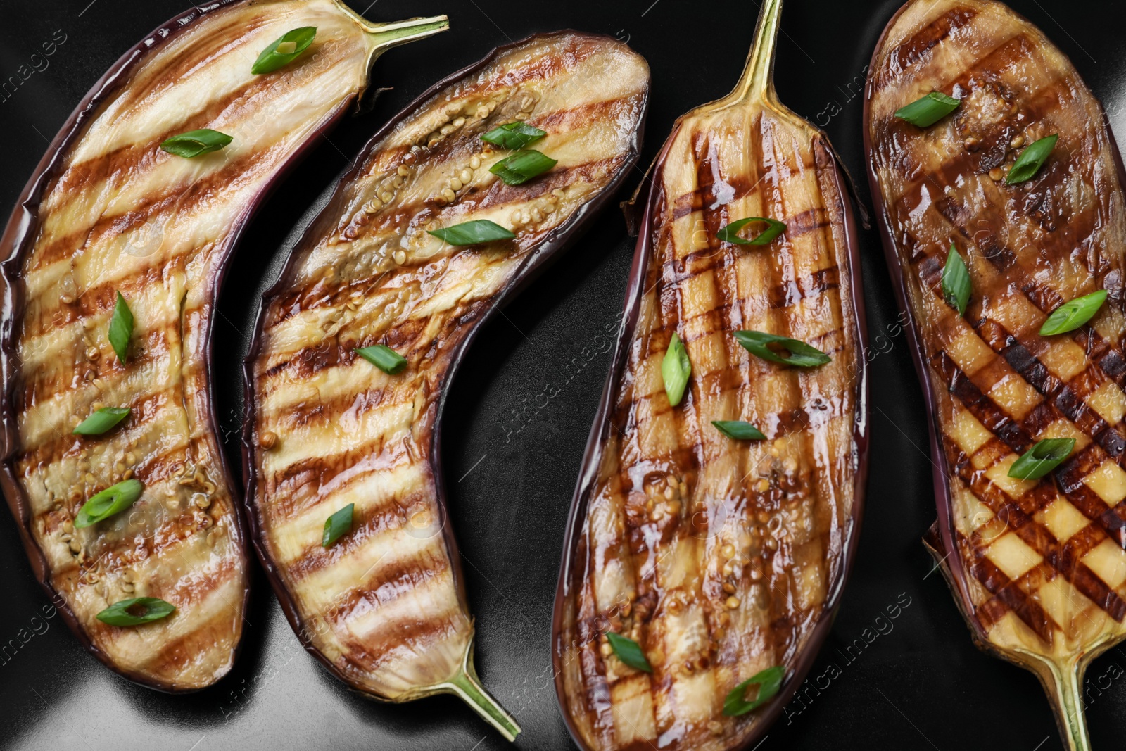 Photo of Delicious grilled eggplant halves with green onion on black plate, closeup